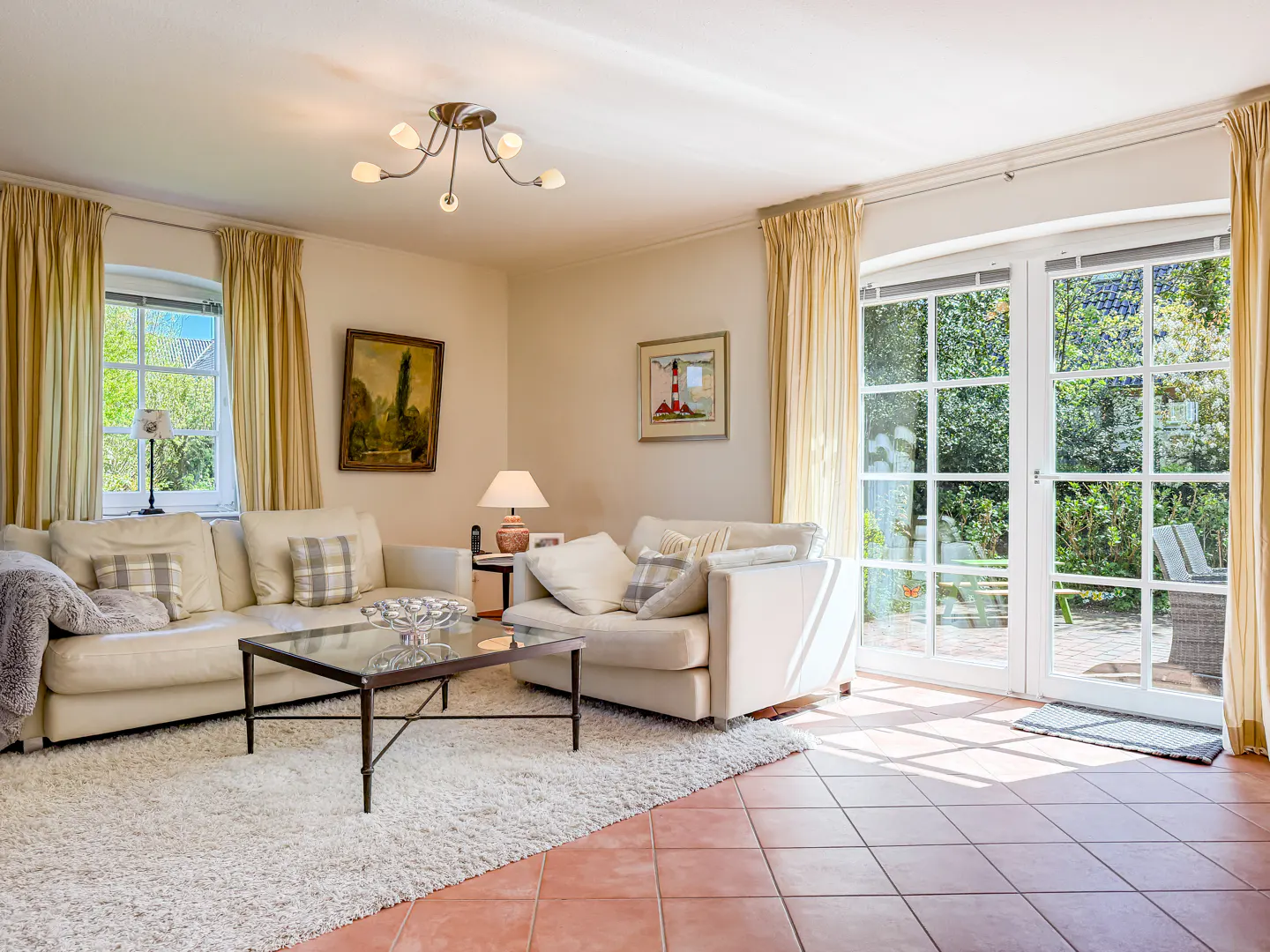 Bright living room with white sofas, a glass coffee table on a white rug, and terracotta tile floors. French doors lead to a green garden.