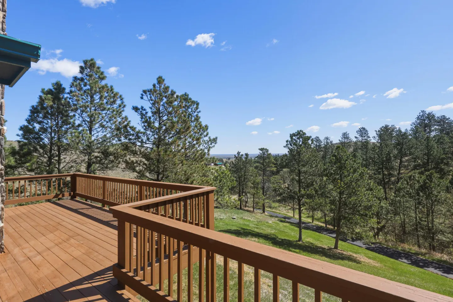 A wooden deck overlooks a green lawn and pine trees under a blue sky with scattered clouds.