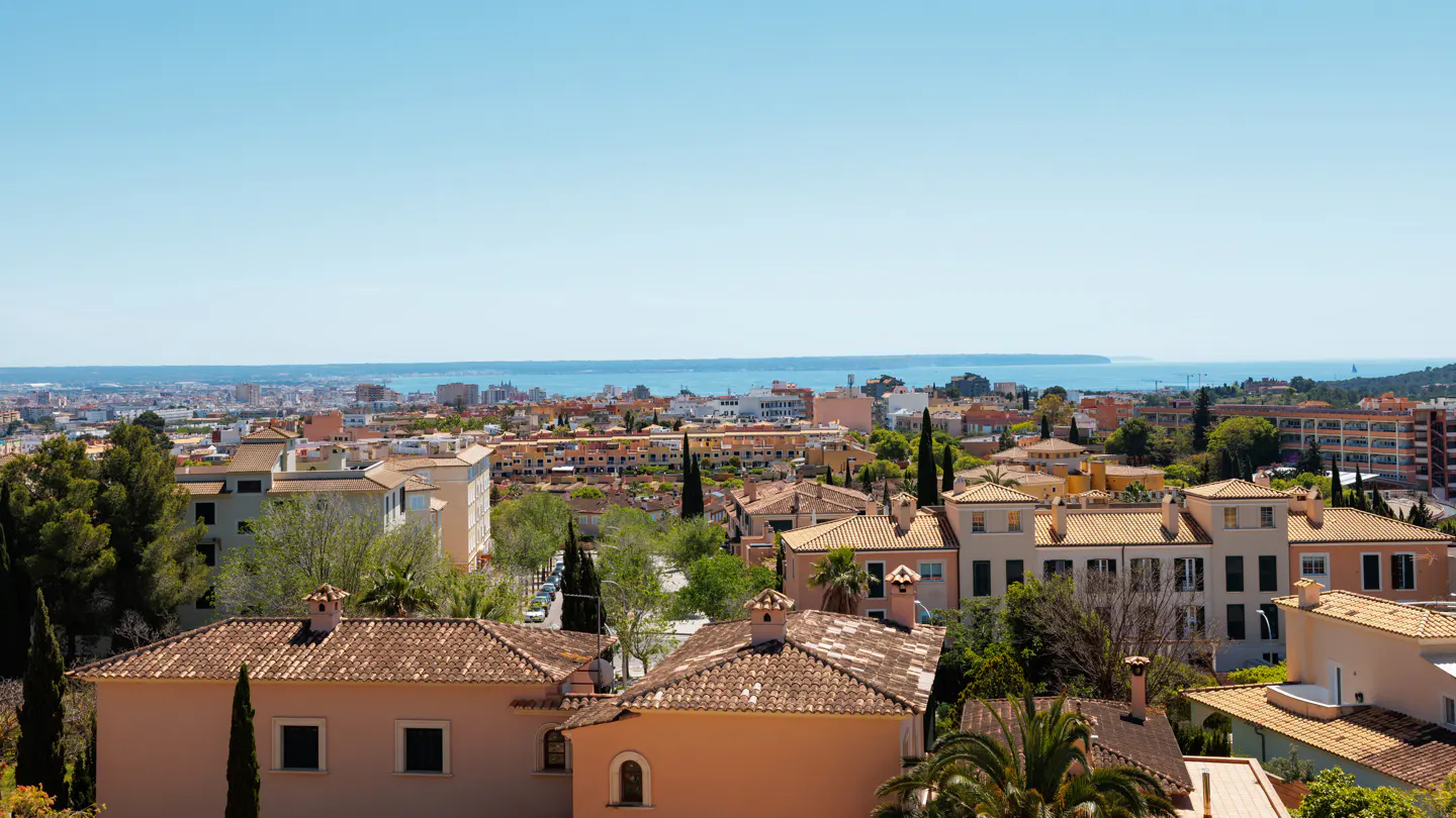 Cityscape view of Mallorca, Spain, with terracotta-roofed buildings, green trees, and the Mediterranean Sea under a clear blue sky.