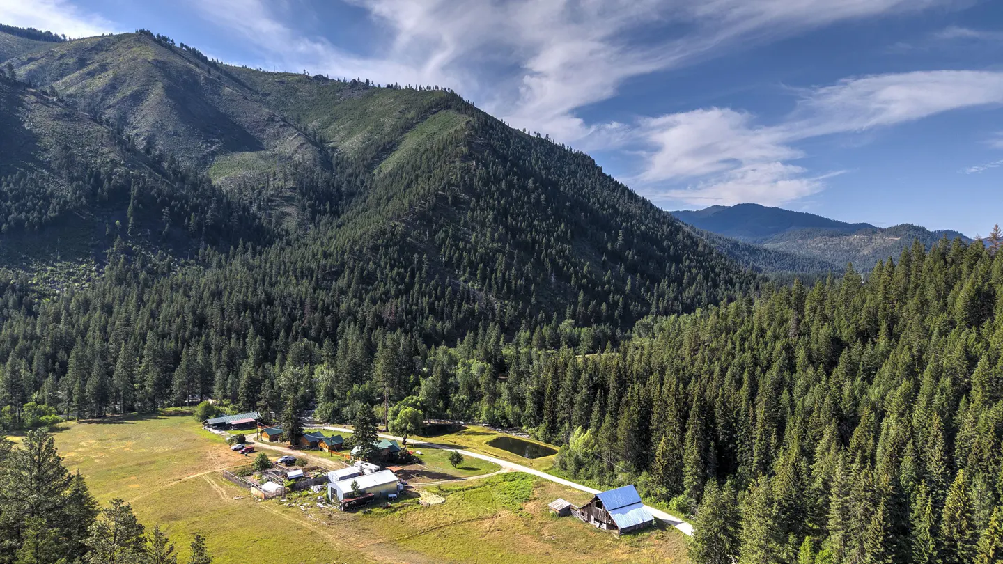 Aerial view of a property with a house, cabins, and barn nestled in a valley surrounded by mountains and a dense forest under a blue sky.