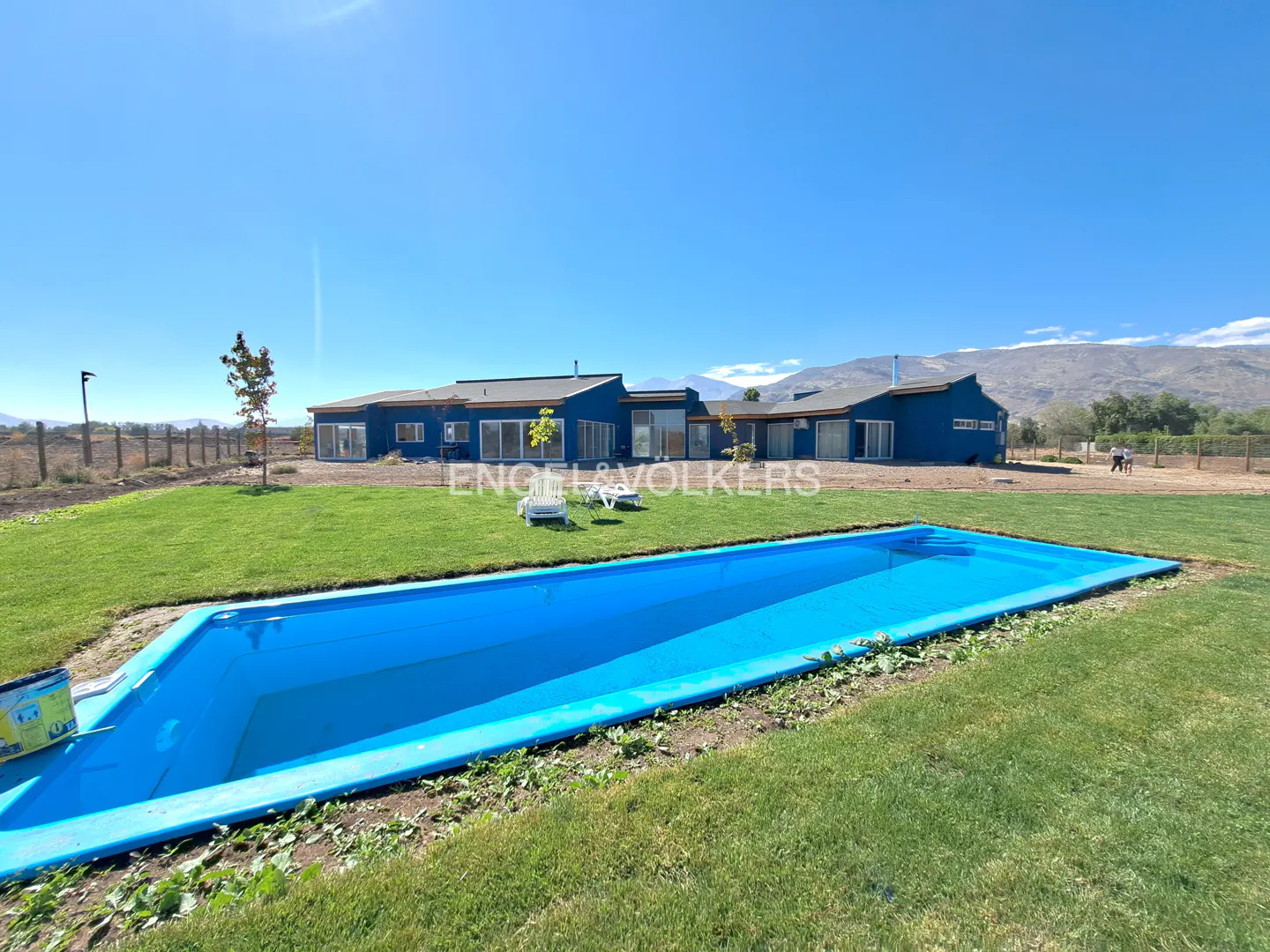 A blue house with a pool and green lawn under a clear blue sky. Mountains are visible in the background.