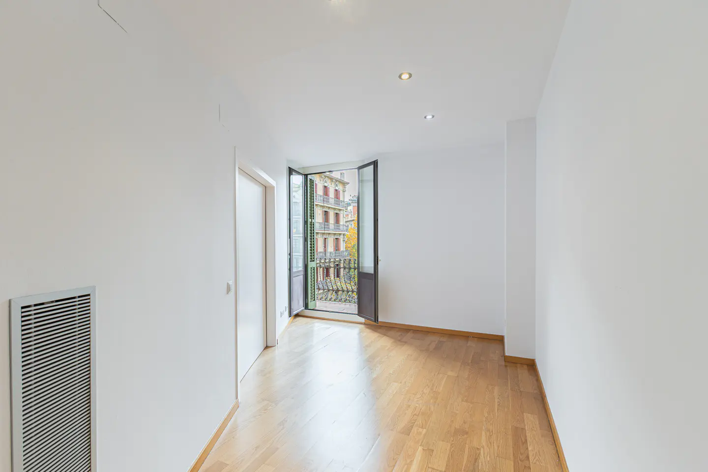 Empty room with white walls, light wood floors, and an open balcony door overlooking a city building.