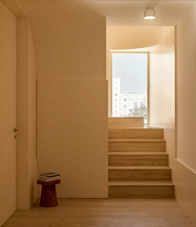 Beige interior with stairs leading to a window. A small stool with books sits near a closed door.