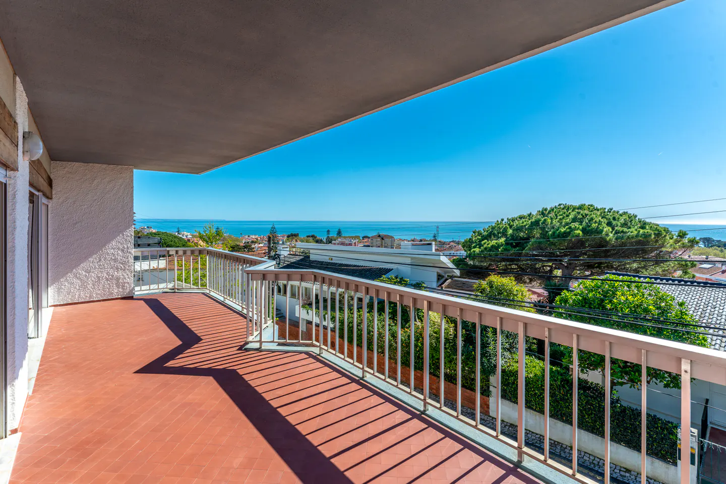 Balcony with red tile floor and white railing overlooks the ocean and a neighborhood with green trees under a clear blue sky.