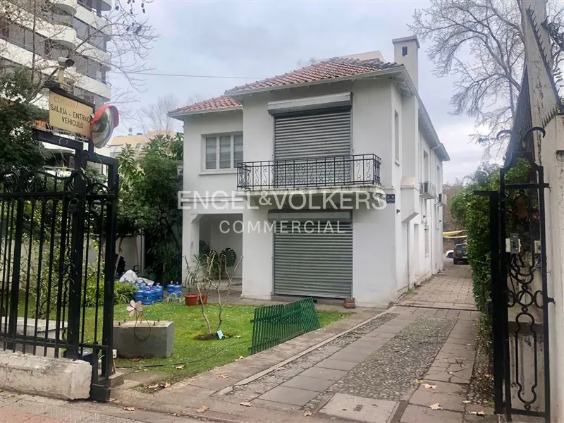 Two-story white building with red tile roof and gray roll-up doors, Engel & Volkers Commercial sign, black iron gate and stone driveway.