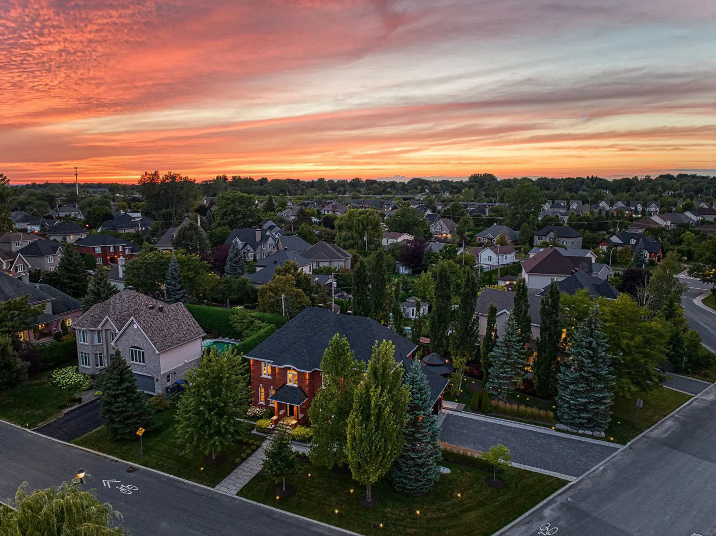 Aerial view of a suburban neighborhood at sunset. Red brick house with black roof, green trees, and orange sky.