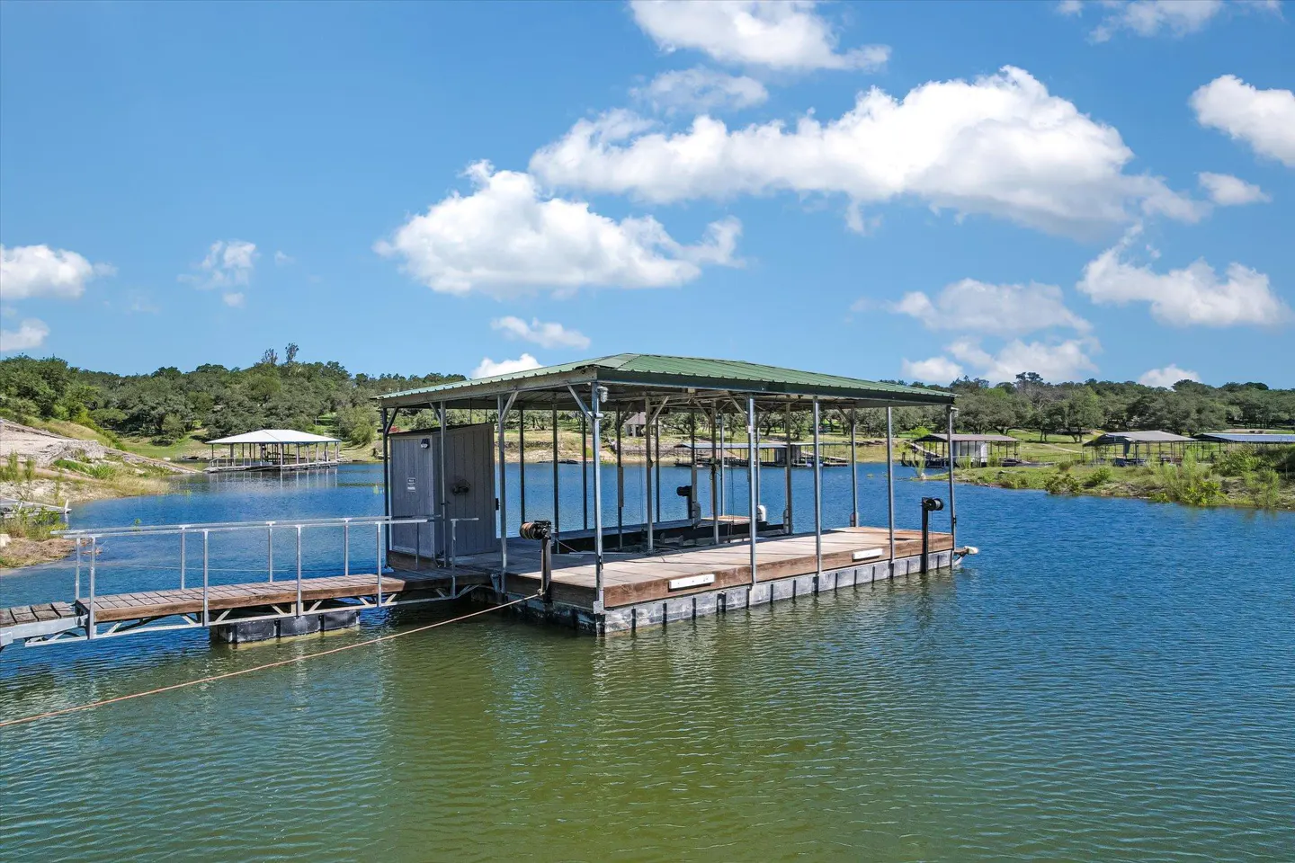 Lake view featuring a covered boat dock with a green roof and a wooden walkway under a blue sky with fluffy white clouds.