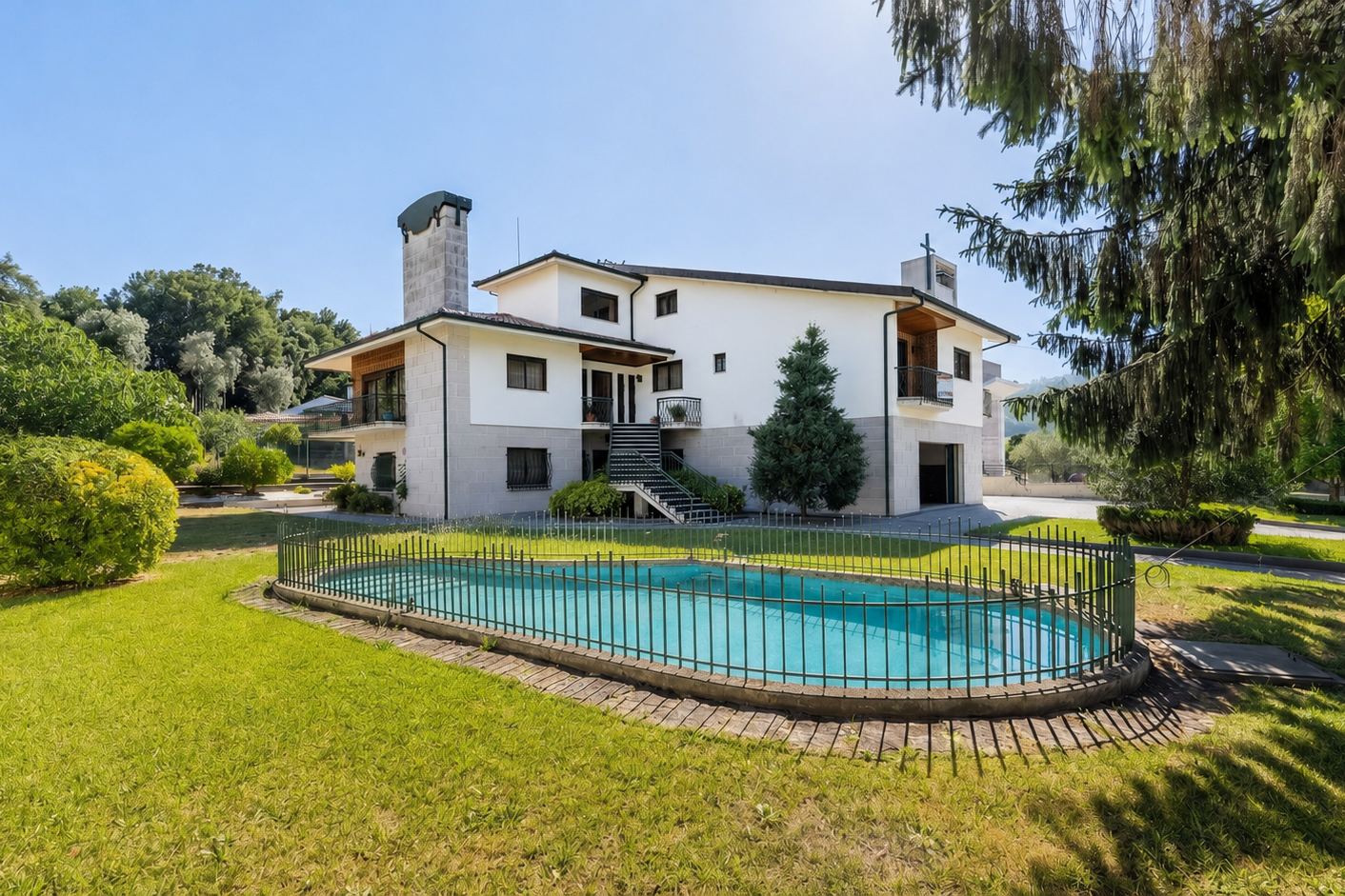 Two-story white house with a stone chimney, a fenced-in swimming pool, and a grassy yard under a clear blue sky.