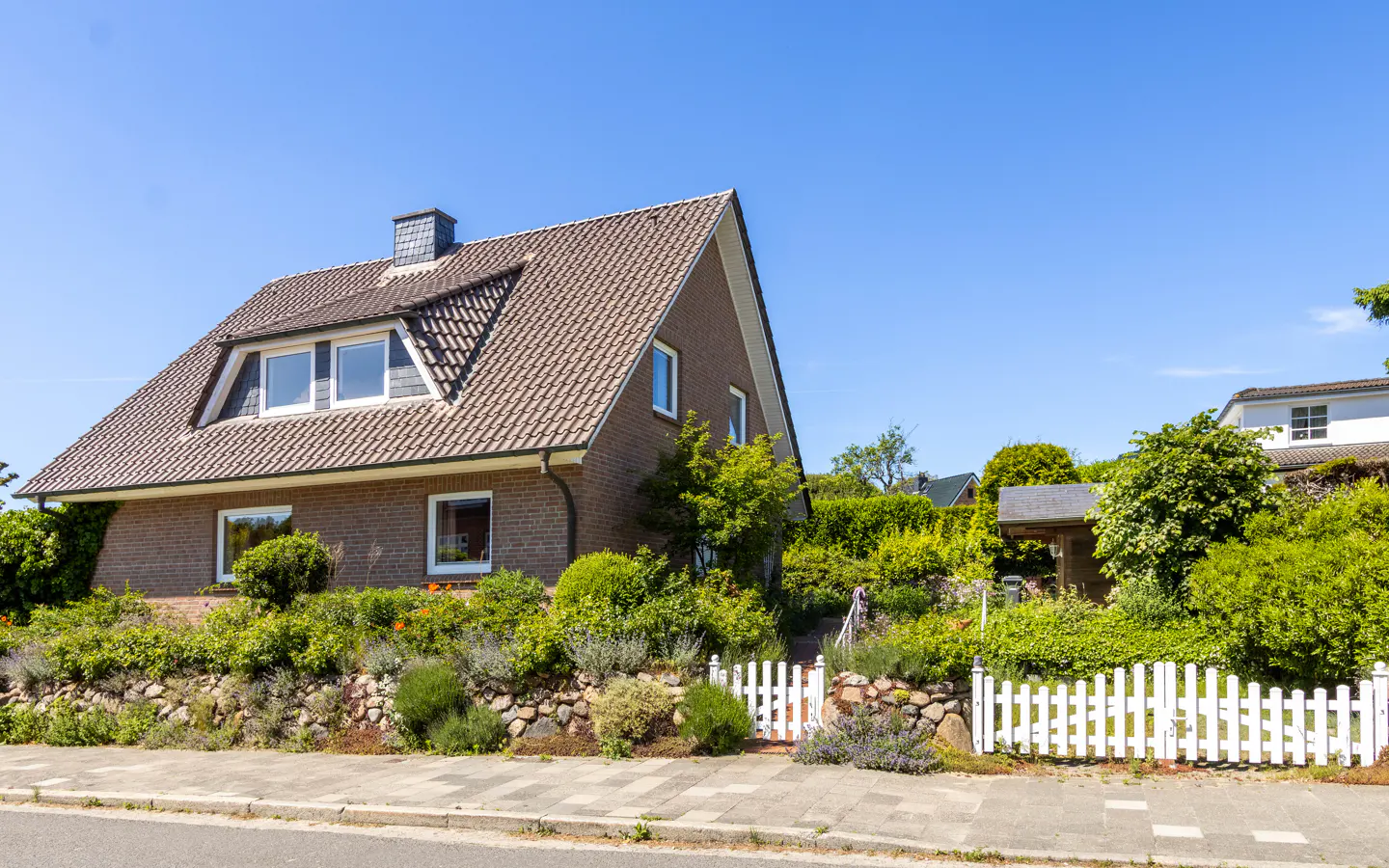 A brown brick house with a tiled roof, surrounded by lush greenery, a stone wall, and a white picket fence under a blue sky.