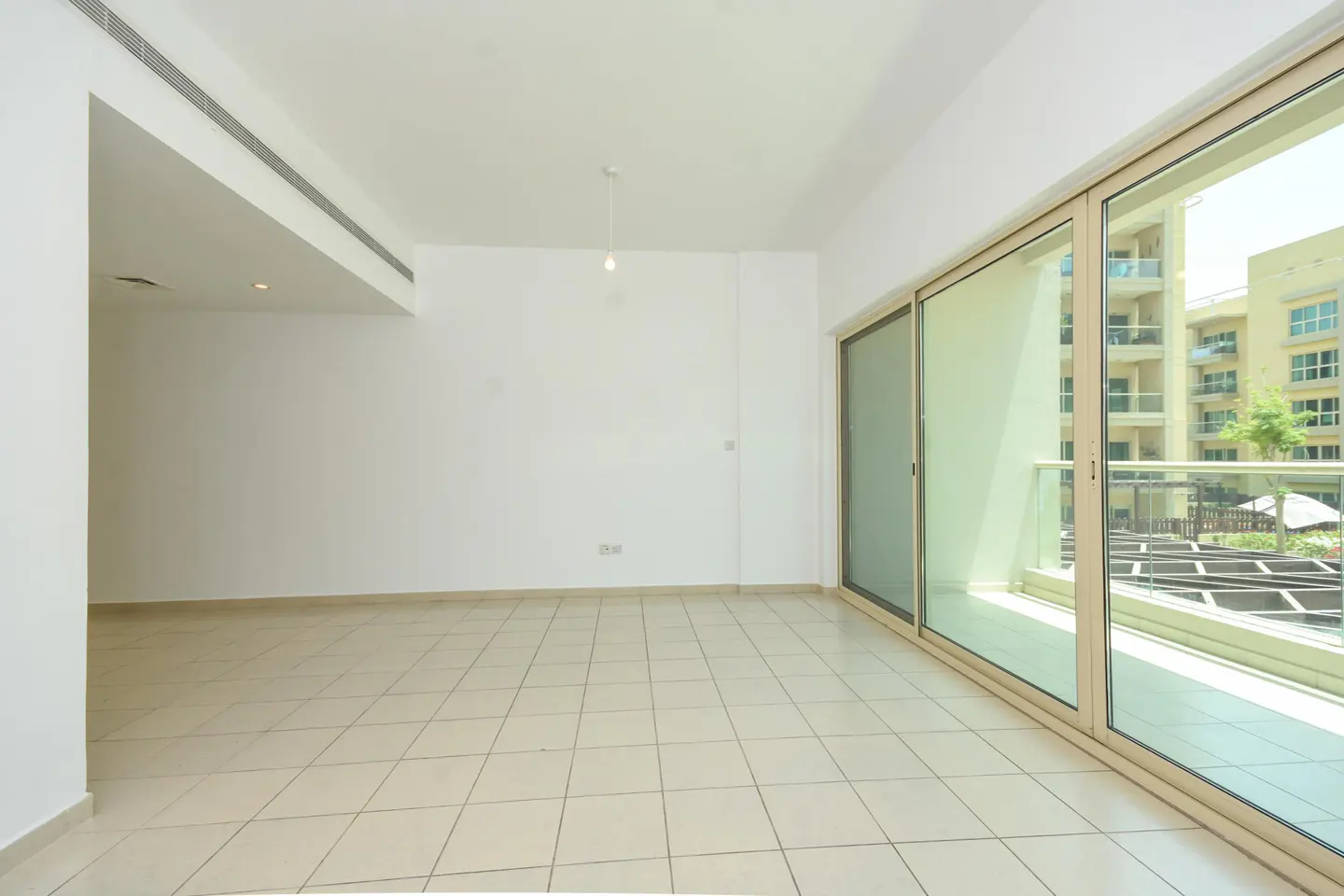 Bright, empty room with white walls and tiled floor. Sliding glass doors lead to a balcony with a view of a building and greenery.