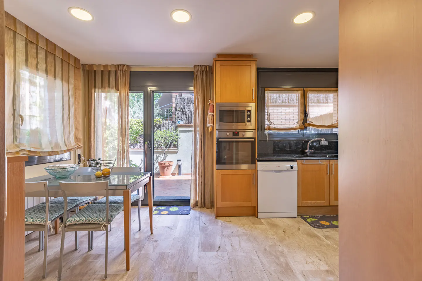 A bright kitchen with a glass table, chairs, and a view to a patio through glass doors. The kitchen features wood cabinets and stainless steel appliances.
