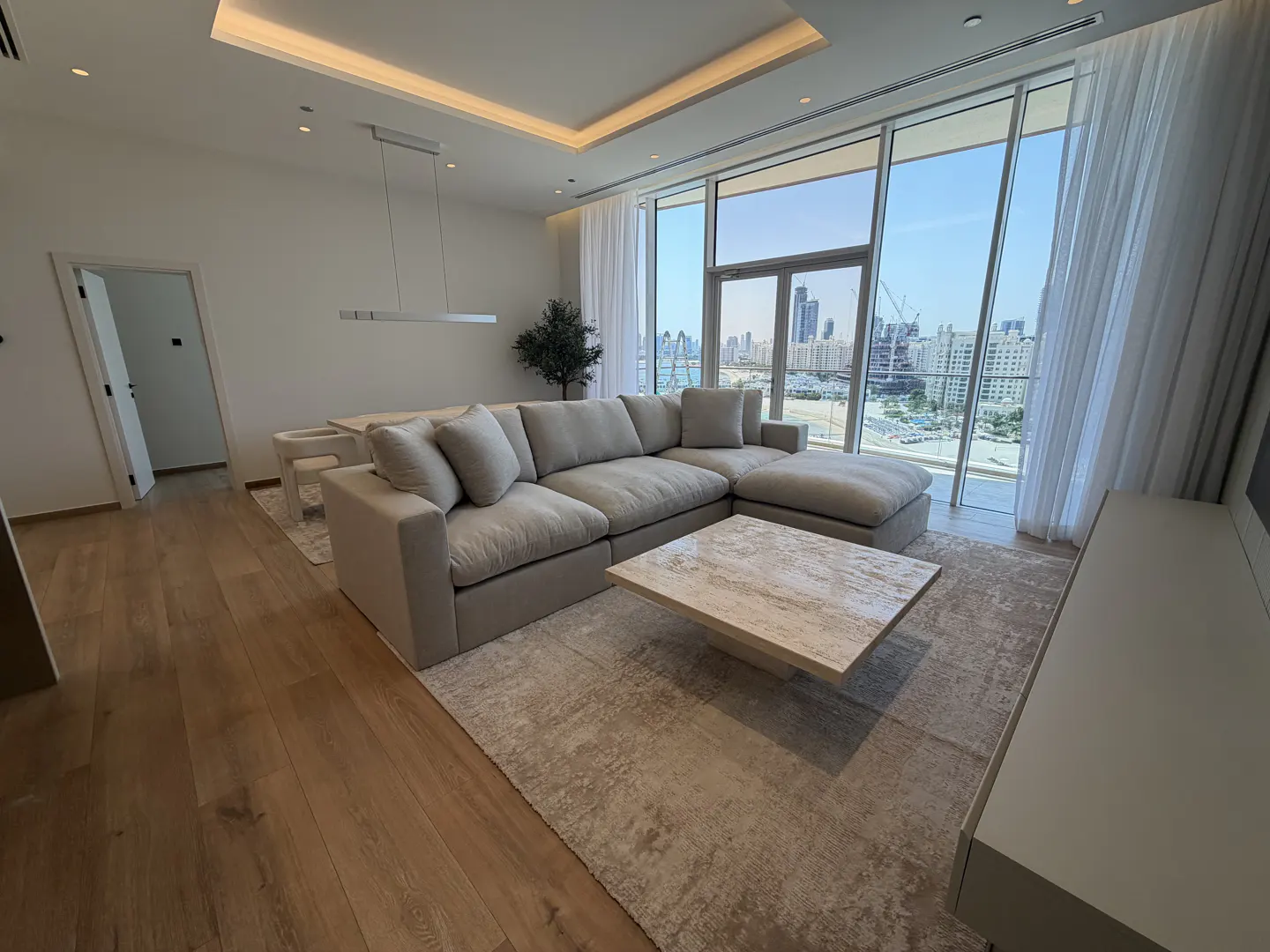 Bright living room with a beige sectional sofa, marble coffee table, and city view through large windows.