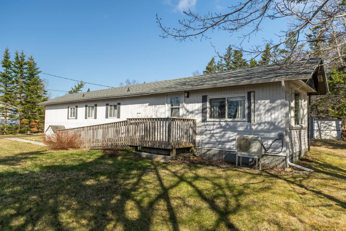Exterior view of a one-story white house with a wooden ramp leading to the front door. Green lawn and trees surround the house.