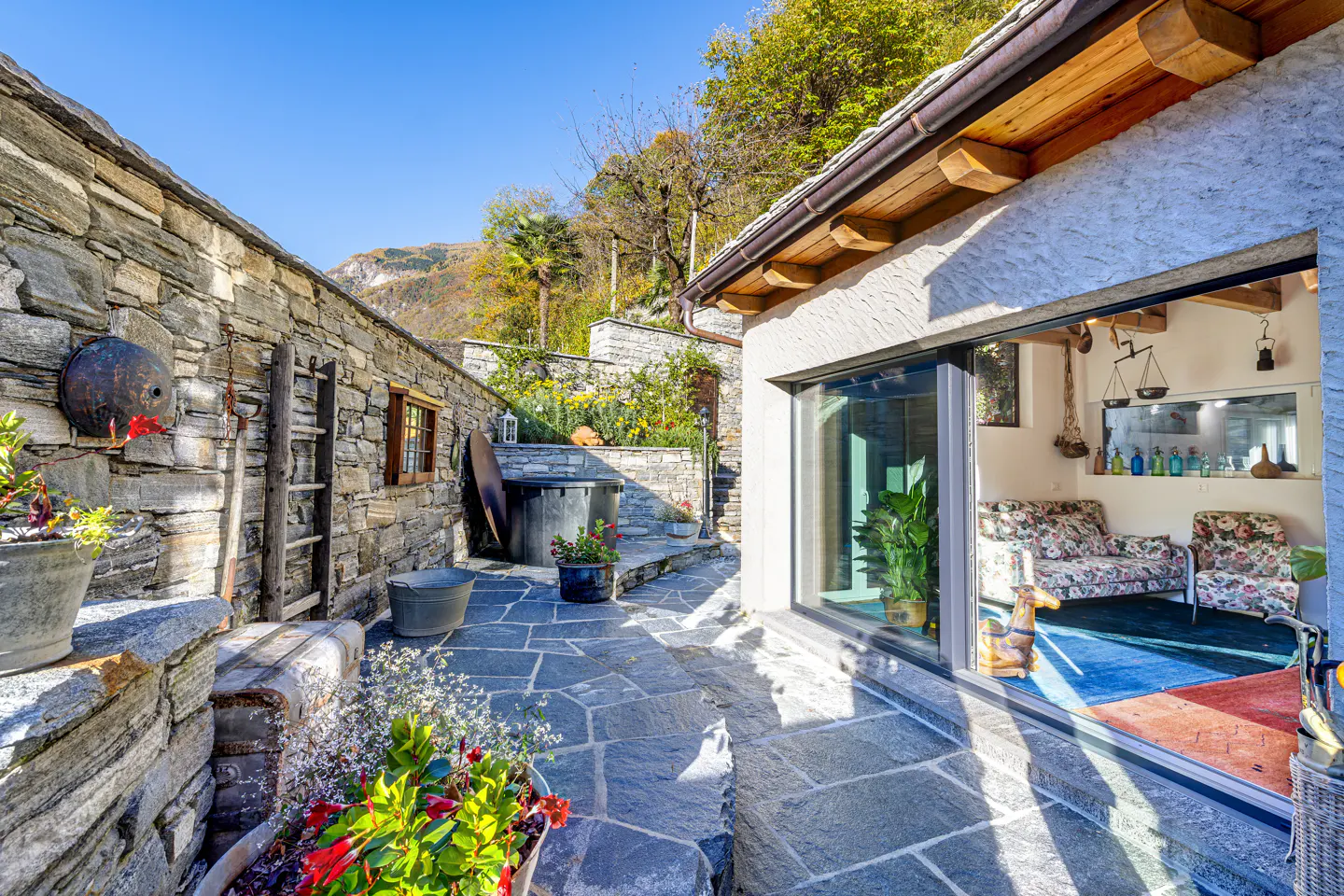 Stone patio with flowers and a stone wall. Inside, floral sofas sit on a blue rug.