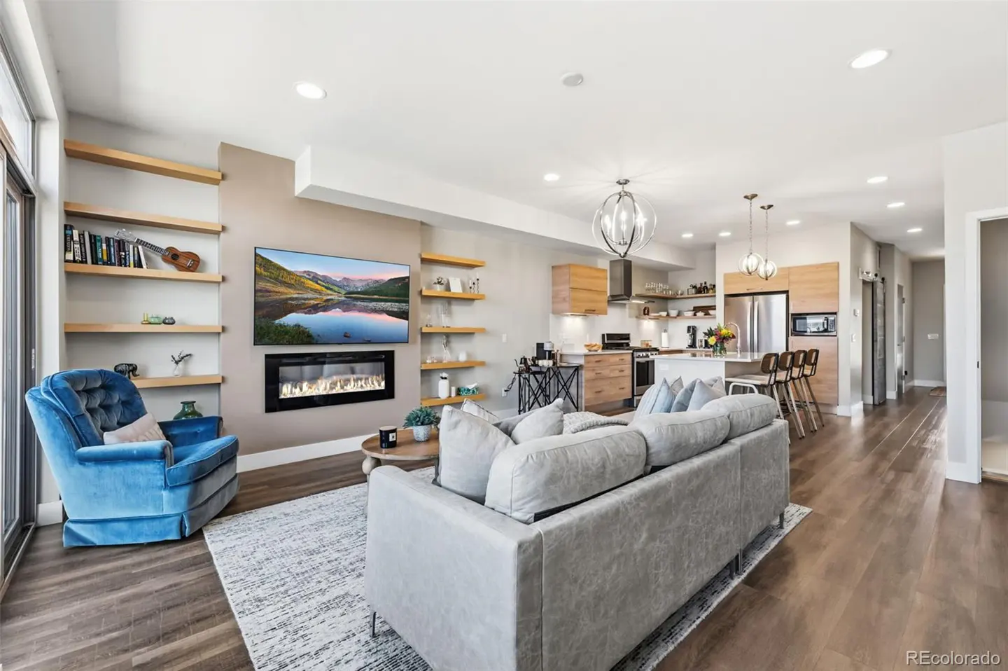 Open-concept living space with a gray sofa, blue armchair, fireplace, TV, and kitchen with wood cabinets and island.