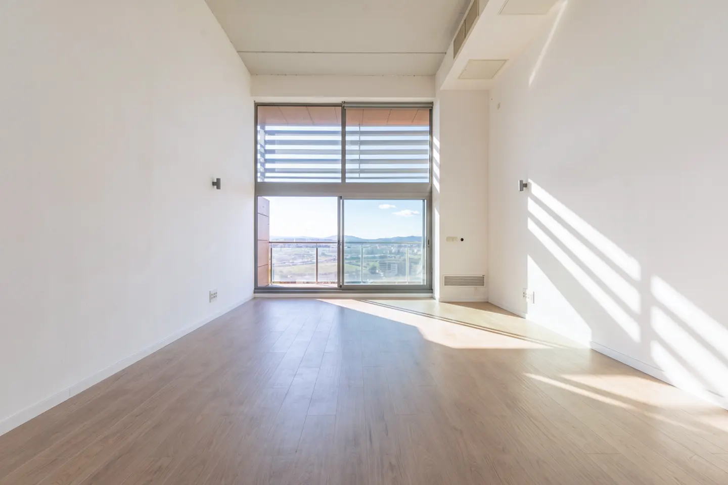 Bright, empty room with wood floors and white walls. Large window with blinds overlooks a city view. Sunlight streams in.