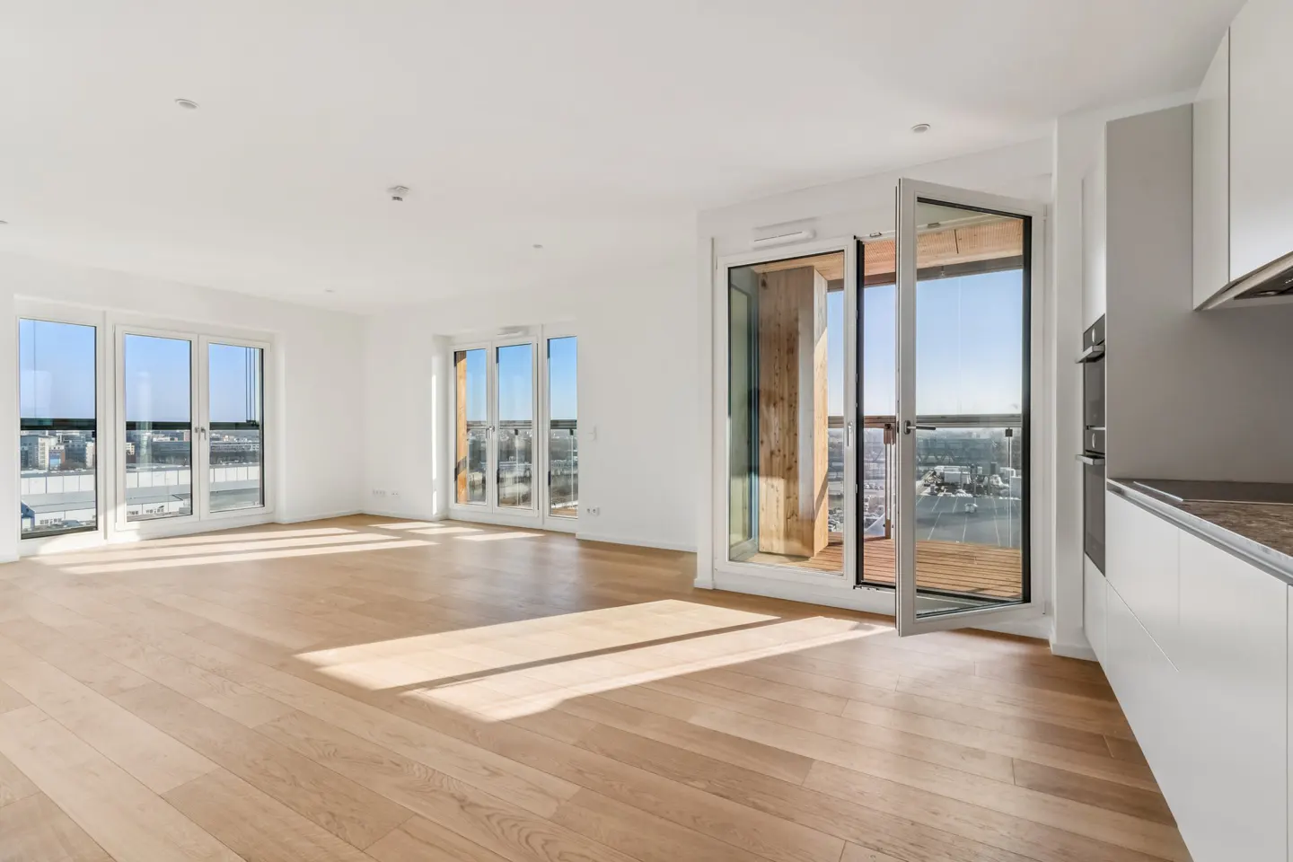Bright, modern apartment interior with hardwood floors, white walls, and large windows offering city views. Open balcony door and kitchen area visible.