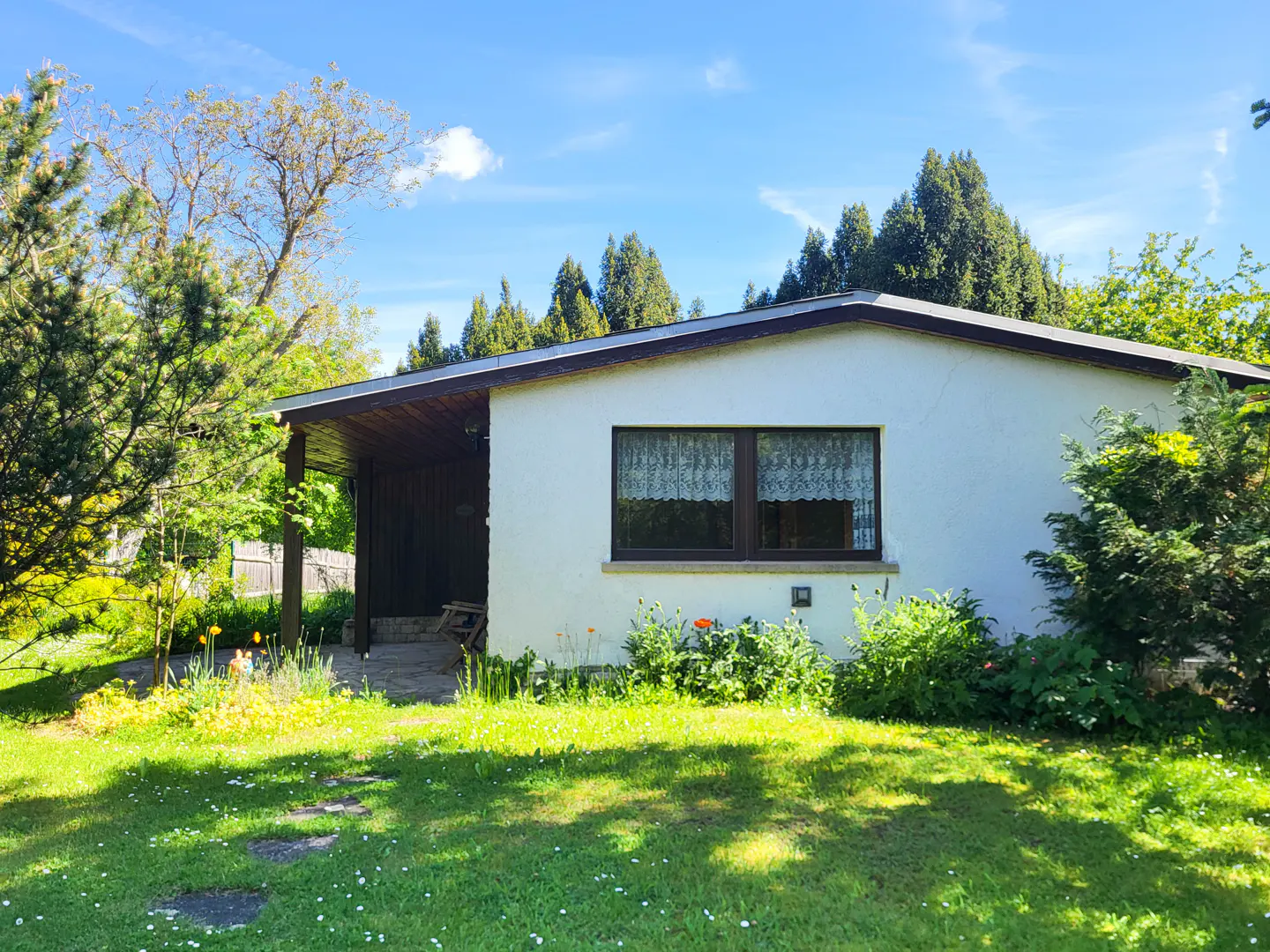 A small, white house with a brown roof and trim, surrounded by green grass, trees, and flowers under a blue sky.
