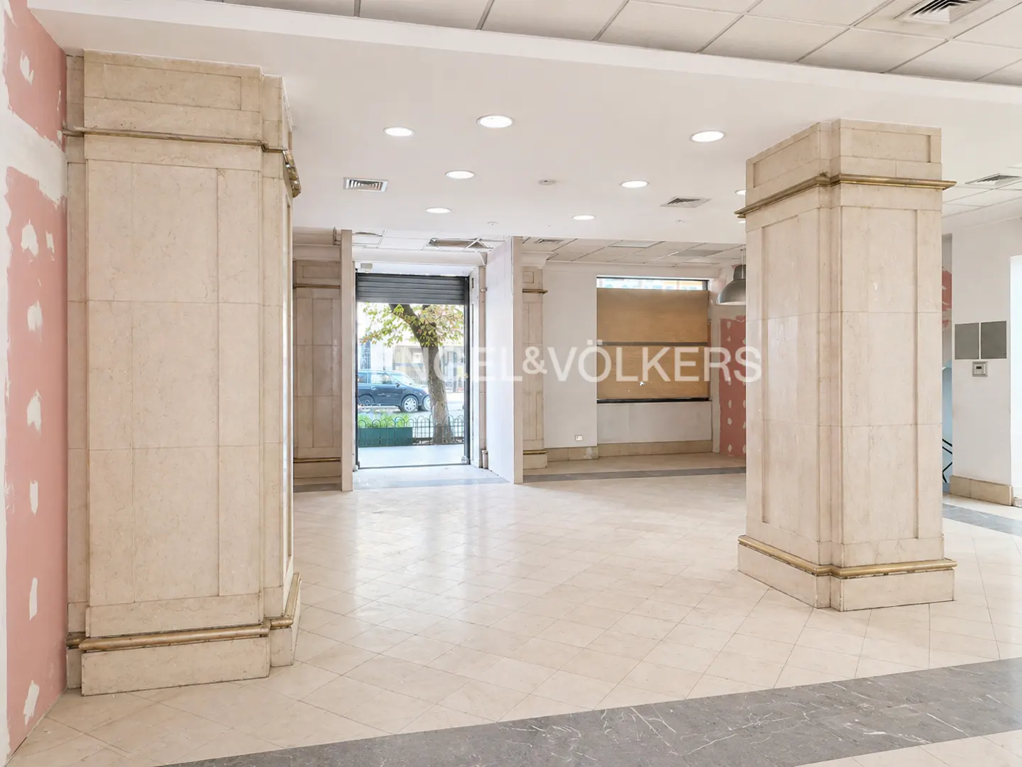 Empty commercial space with beige tile floors, large columns, and an open storefront with a view of the street.