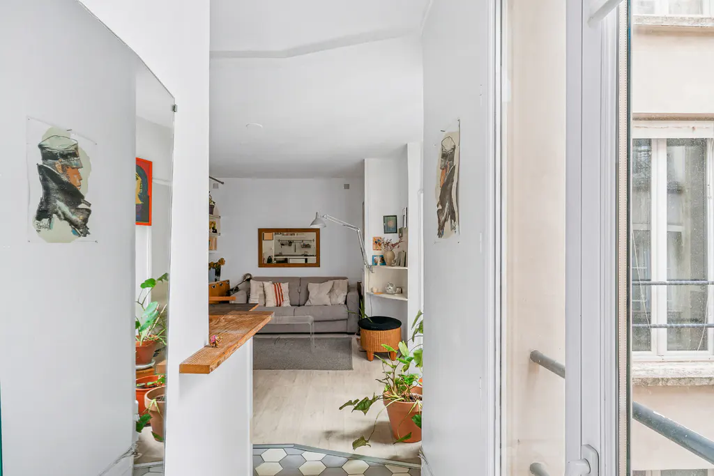 View into a bright apartment with white walls, art, plants, and a gray sofa. A window shows a building exterior.