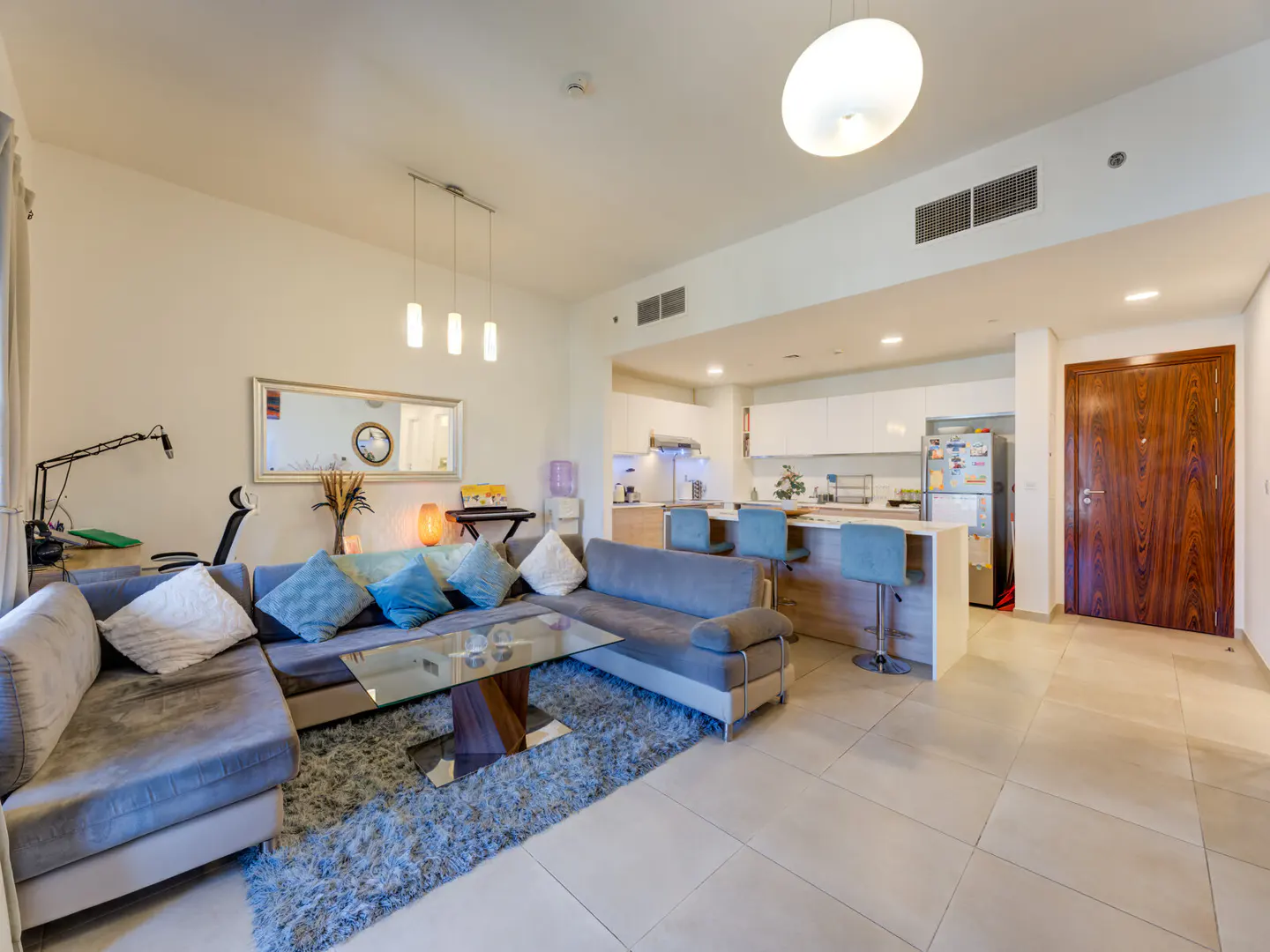 Open-concept living space with a gray sectional sofa, glass coffee table, and kitchen island with blue stools.