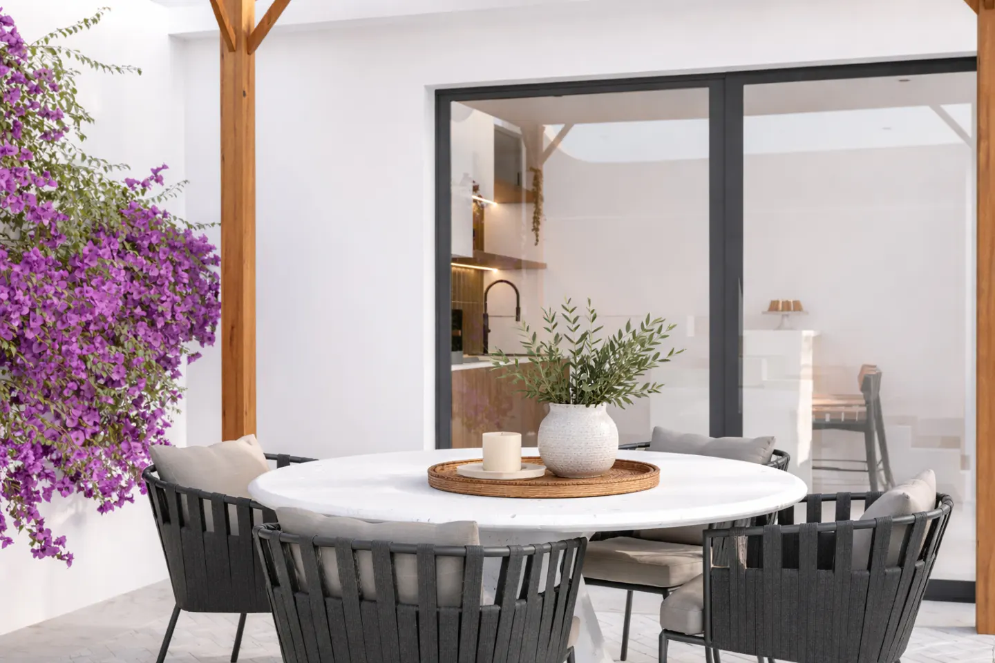Outdoor patio with a white table, gray chairs, and purple bougainvillea. A vase and candle sit on the table. Sliding glass doors lead to the kitchen.