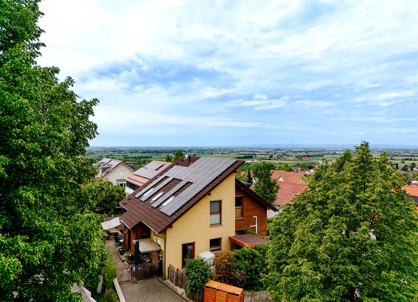 High-angle view of a yellow house with solar panels on its roof, surrounded by lush green trees and a distant landscape.