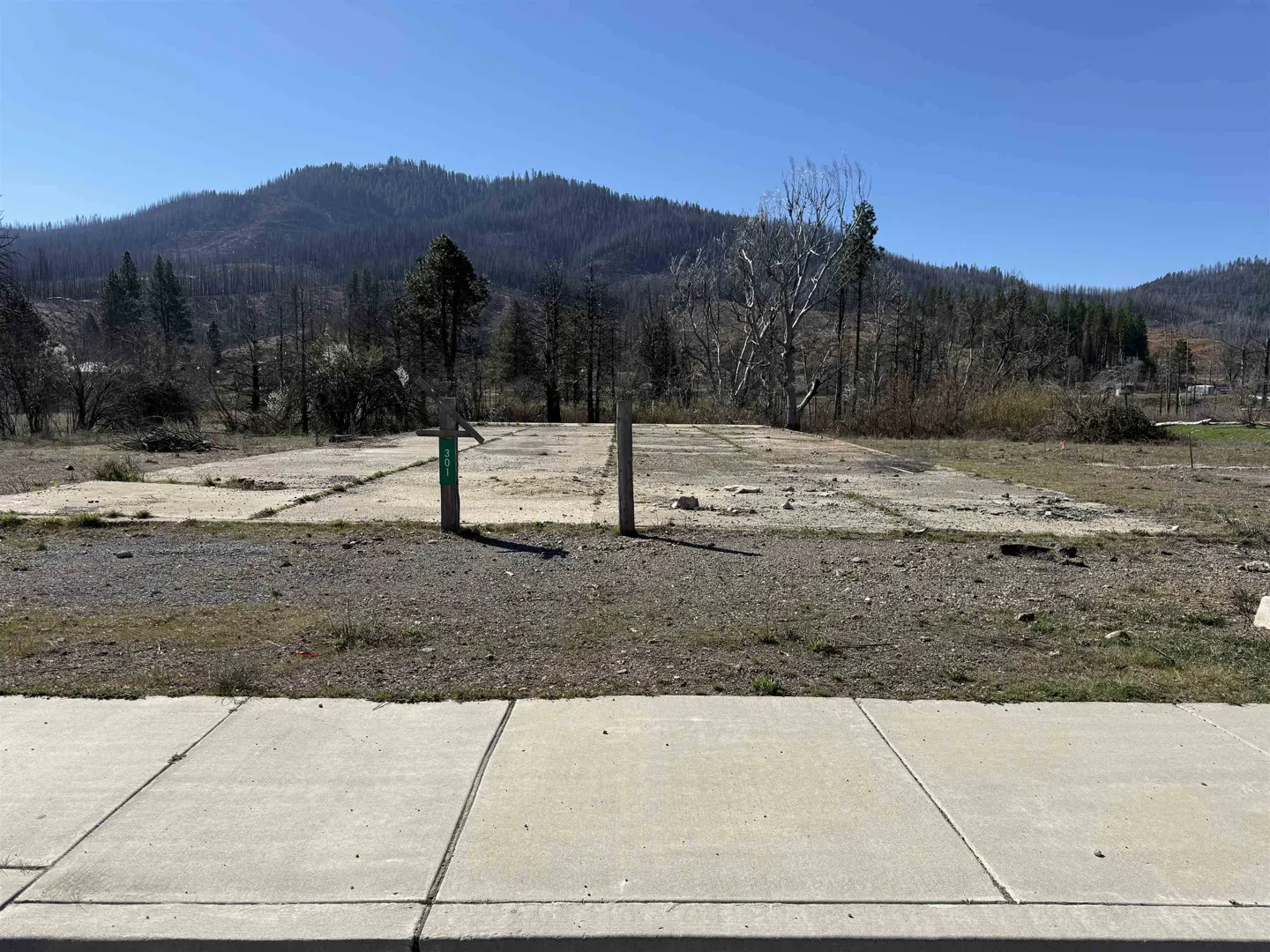 A vacant lot with a concrete foundation slab, gravel, and a mountain backdrop under a clear blue sky.