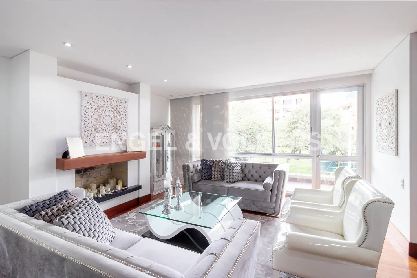 Bright living room with white walls, grey sofas, white chairs, and a glass coffee table. A fireplace and large windows add to the room's appeal.