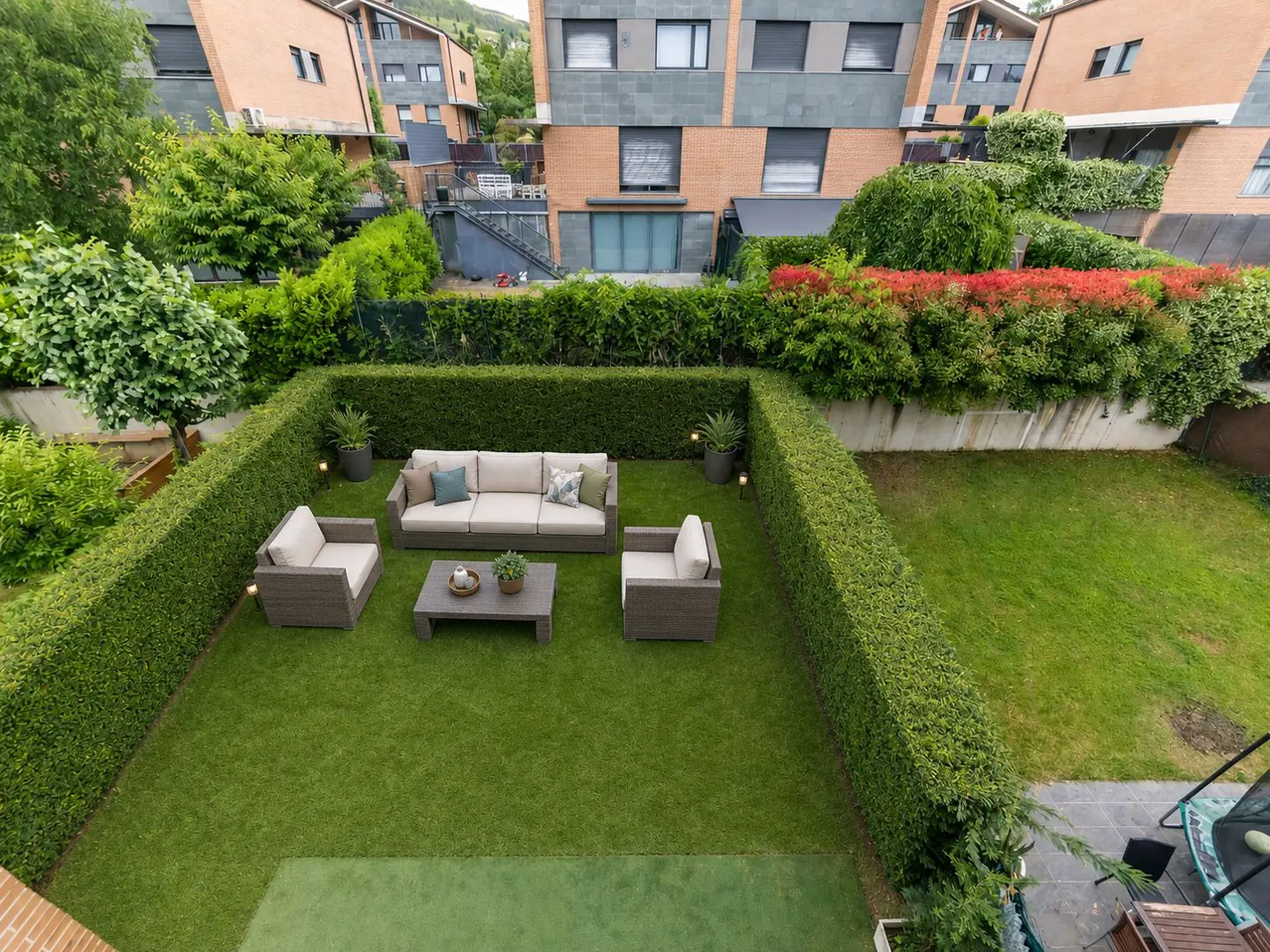 Aerial view of a manicured backyard with a beige sofa, chairs, and coffee table on green grass, surrounded by tall, green hedges. Brick buildings are in the background.