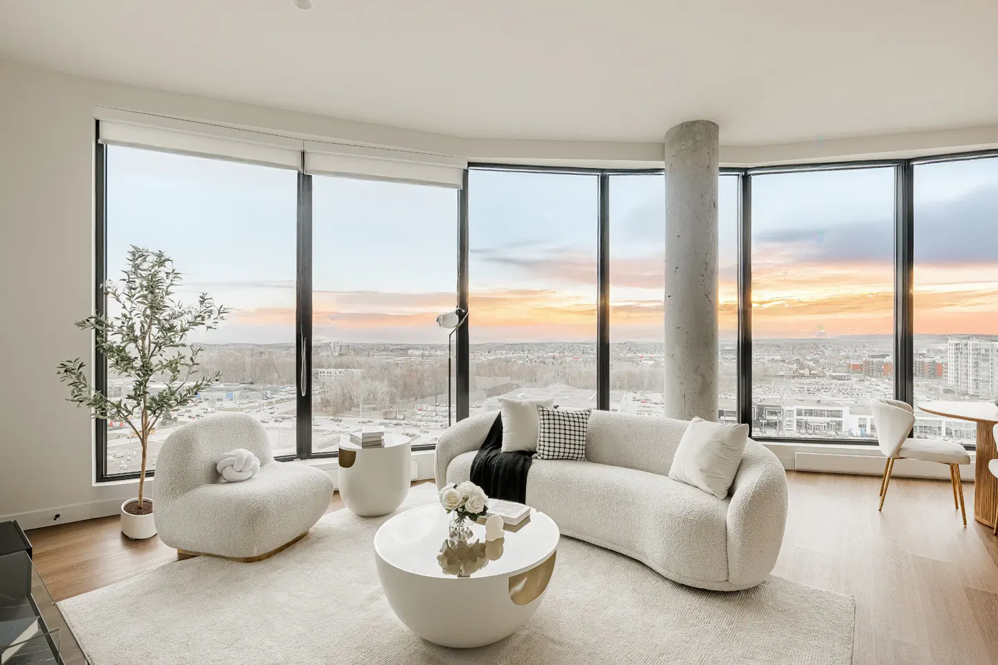 Bright, modern living room with floor-to-ceiling windows showing a sunset view. White, curved sofa and chair, round coffee table, and wood floors.