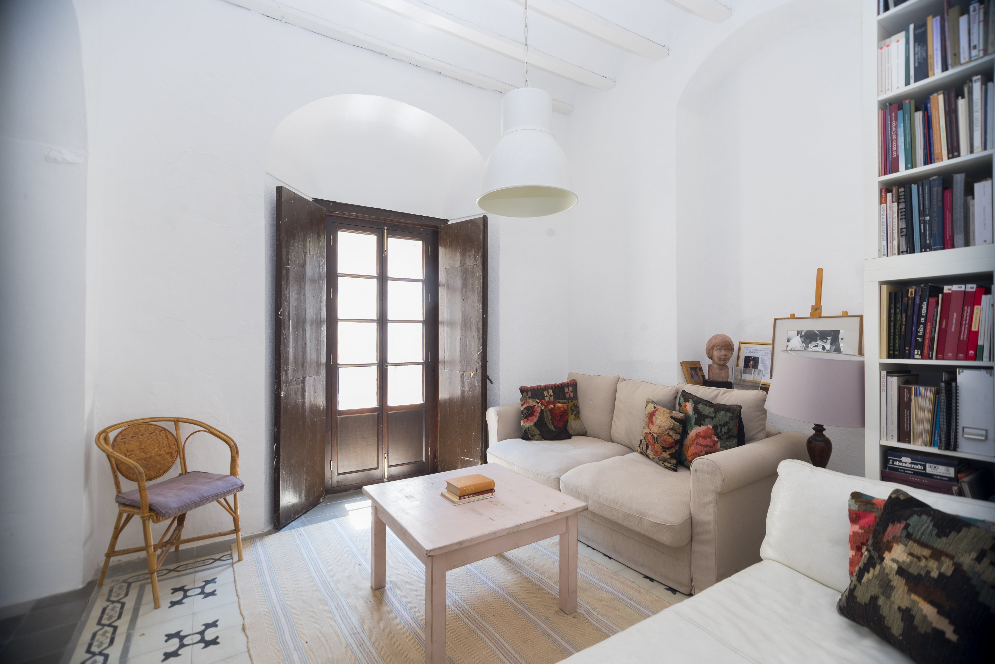 Bright living room with white walls, beige sofas, a light pink coffee table, a rattan chair, and a tall bookshelf.