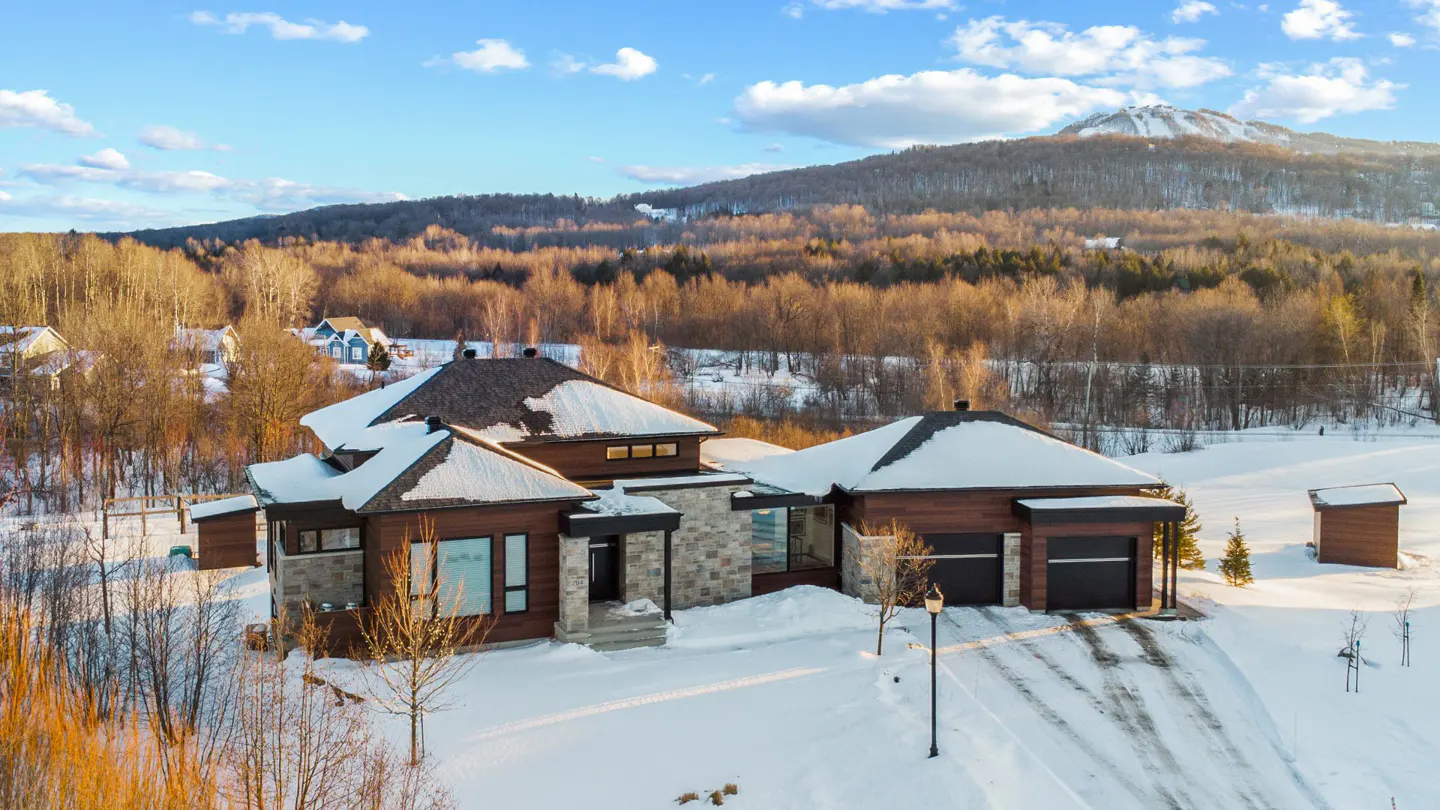 A modern brown house with a dark roof is surrounded by snow, with a mountain backdrop.