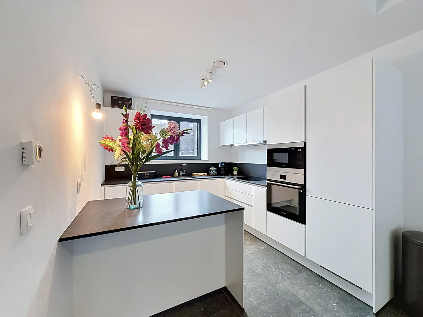 Modern white kitchen with a black island, dark countertops, built-in oven, and a vase of colorful flowers.