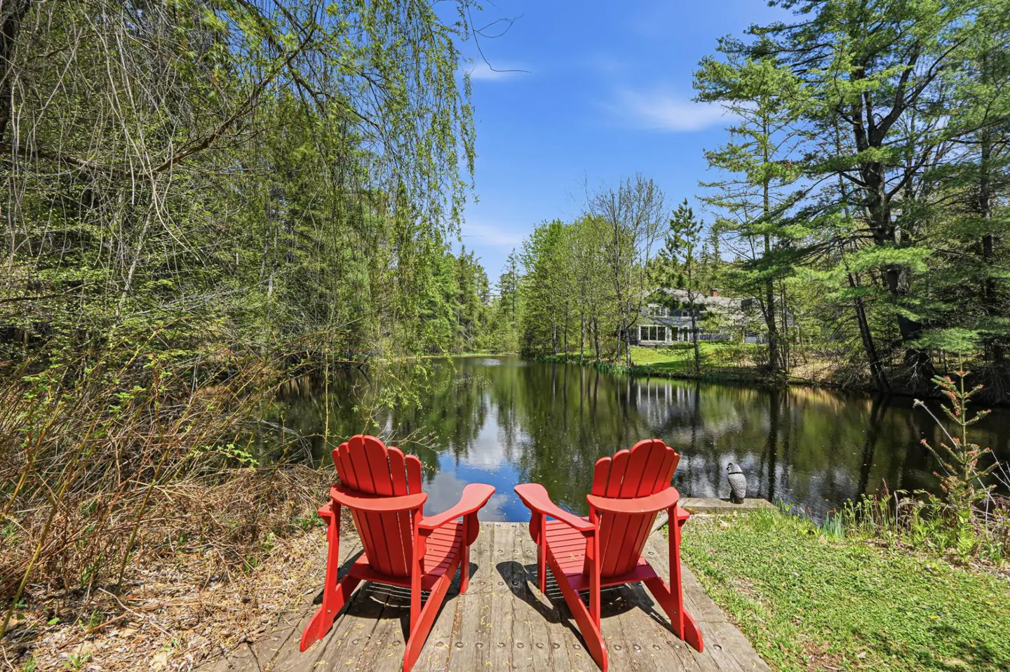 Two red Adirondack chairs sit on a wooden dock overlooking a calm lake surrounded by green trees and a house in the distance.