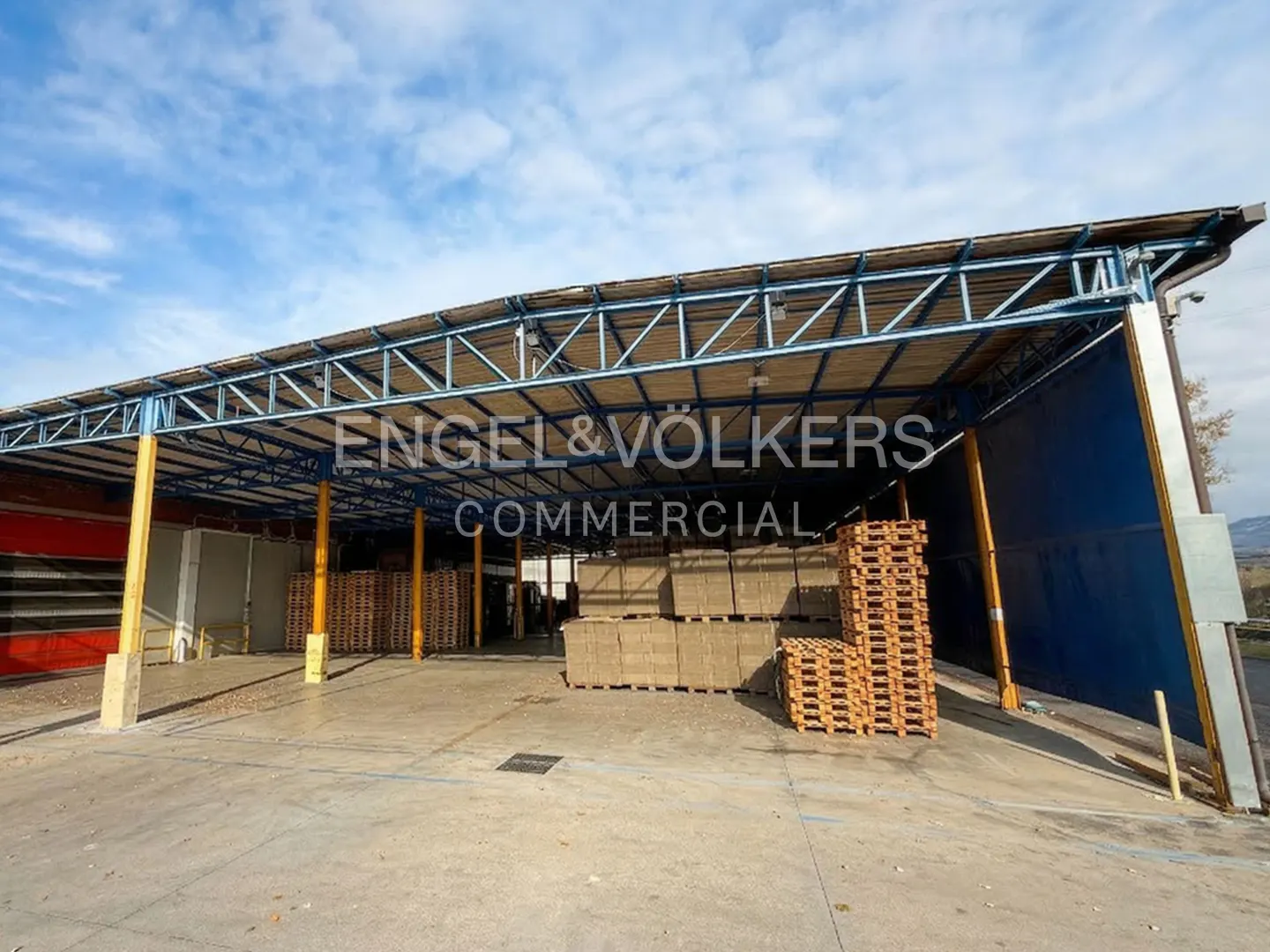 Commercial property with a blue metal awning, stacks of pallets, and the Engel & Völkers Commercial logo.