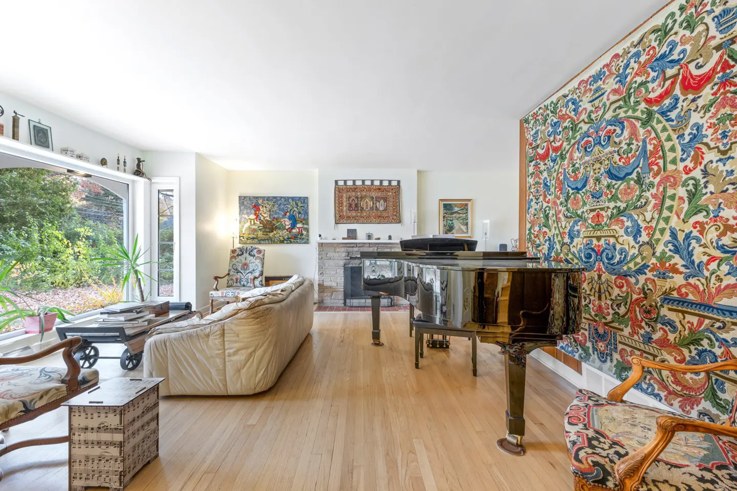 Bright living room with hardwood floors, a grand piano, and a large tapestry on the wall. A sofa and chairs provide seating.