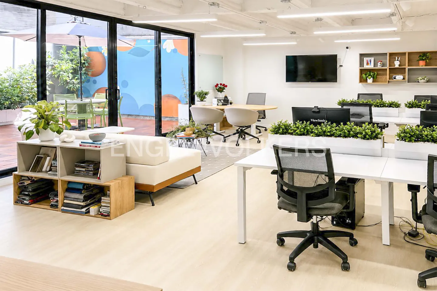Bright, modern office space with white desks, black chairs, and greenery. A lounge area features a cream sofa and open shelving. Large windows offer outdoor views.