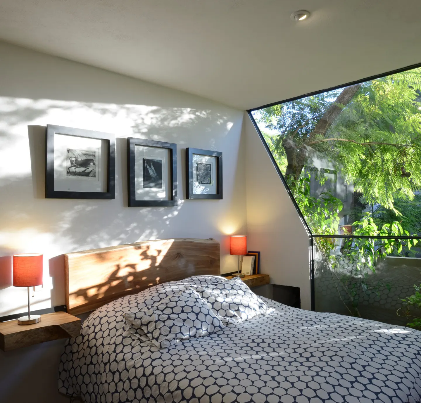 Bedroom with a wood headboard, patterned bedding, and three framed pictures on the wall. A large, angled window shows green trees outside.