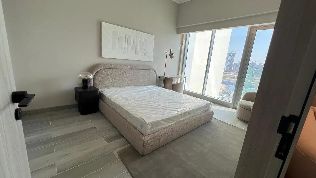 A modern bedroom with a beige bed, a black nightstand, and a large window with a city view.