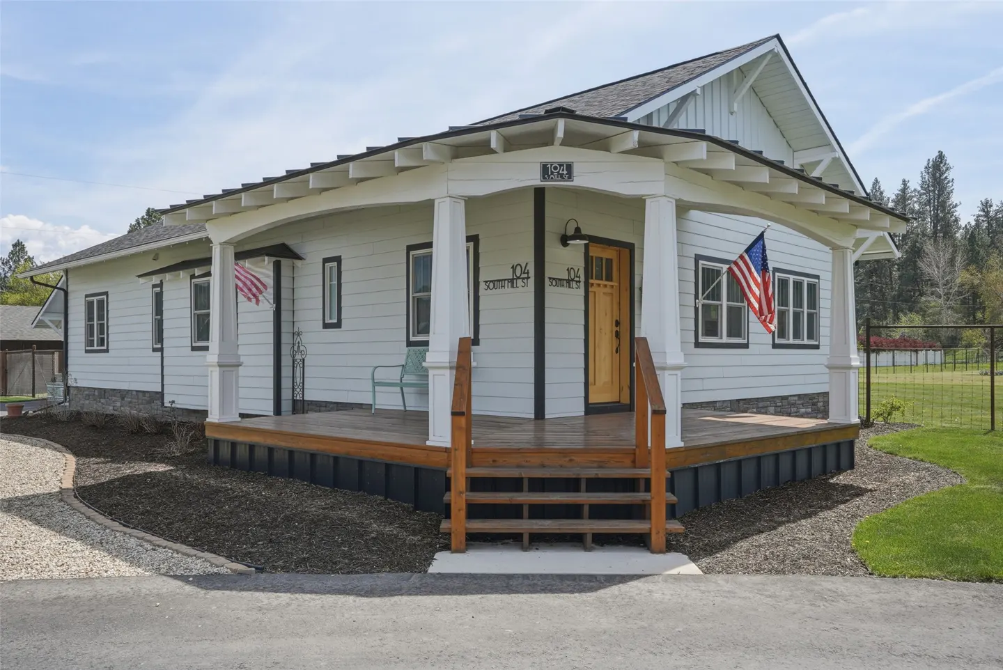 Exterior of a white, one-story home with a wooden porch, American flags, and black trim.