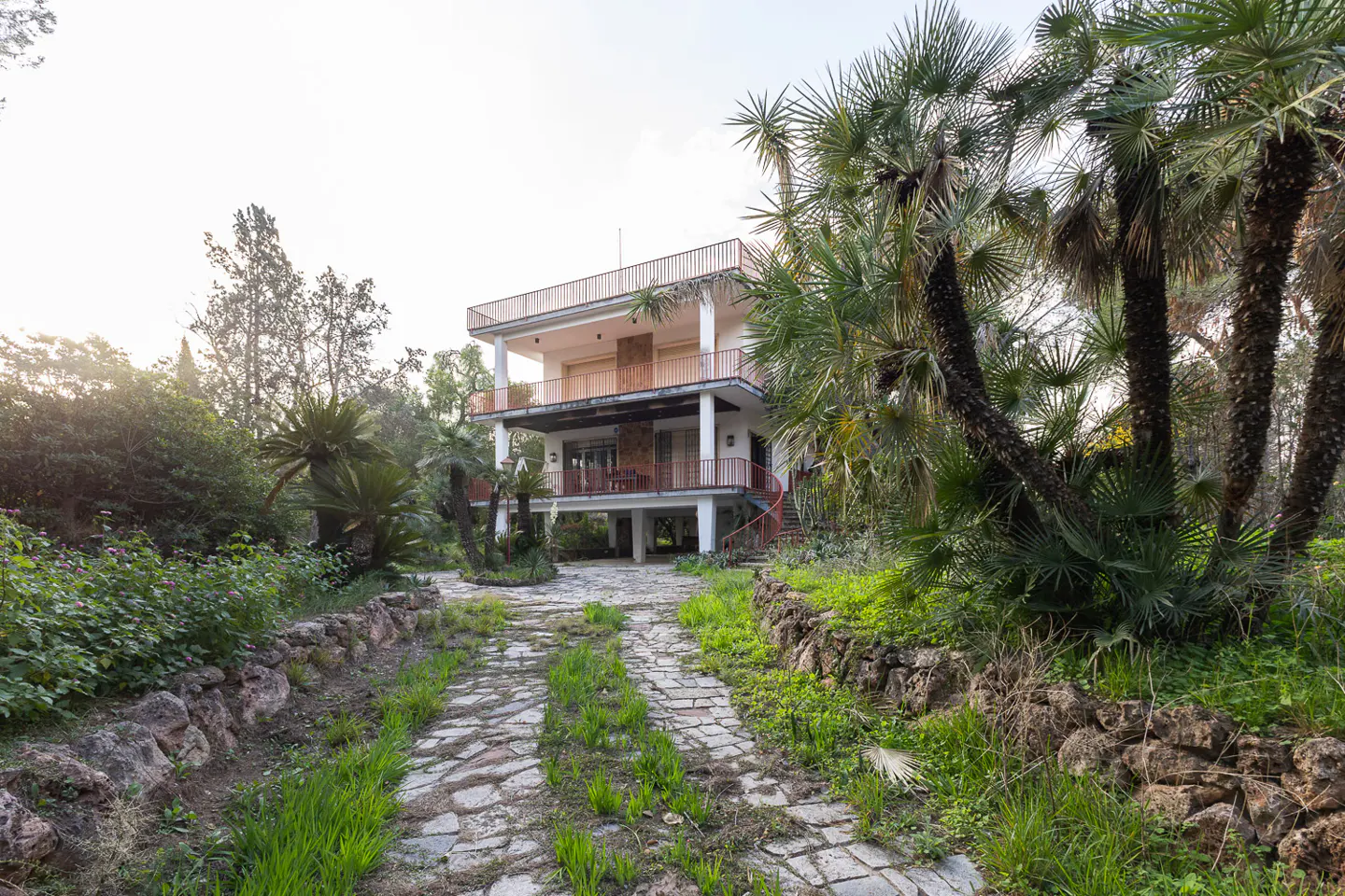 Two-story white house with red railings, surrounded by palm trees and greenery. A stone path leads to the house.