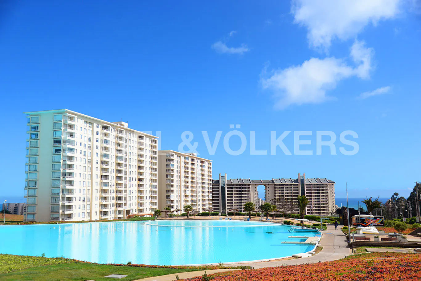Exterior view of white condo buildings with balconies, a large turquoise pool, and a clear blue sky.