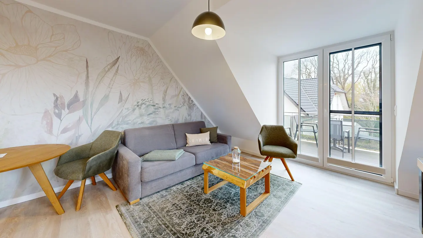A cozy living room with a gray sofa, olive chairs, and a wooden table on a patterned rug. A floral mural adorns the wall. Balcony doors let in natural light.