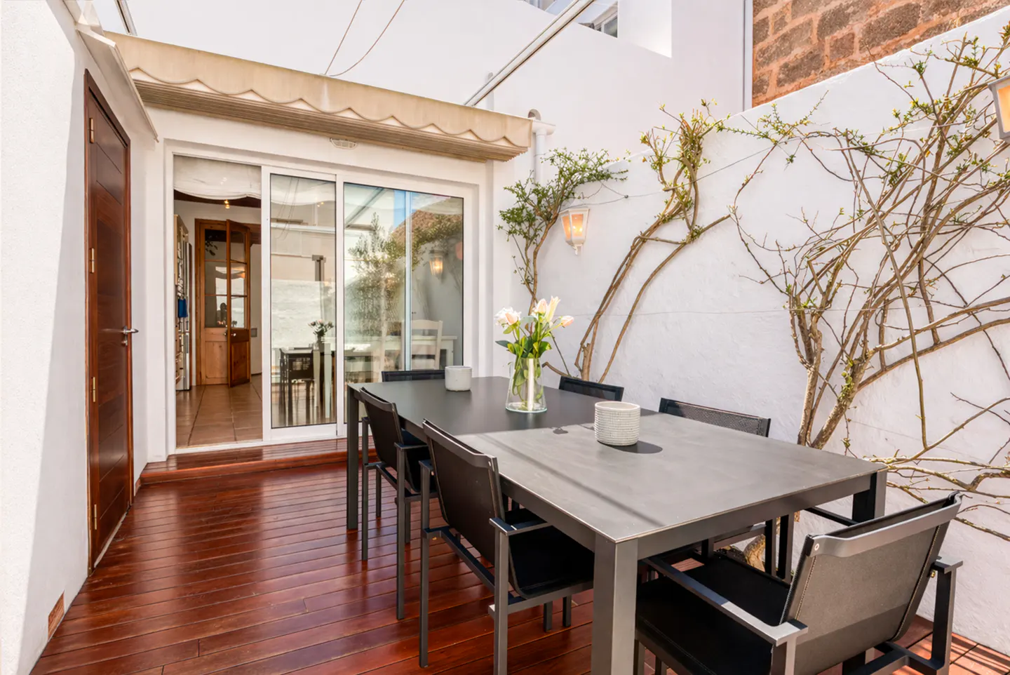 Outdoor patio with a dark table and chairs on a wood floor. White walls with climbing vines and a sliding glass door to the interior.