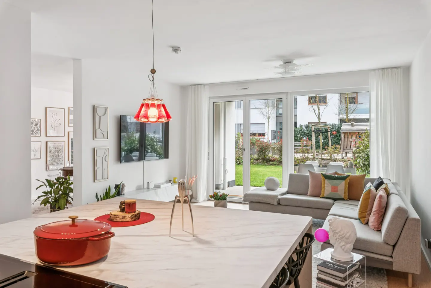 Bright, modern living space with a marble island, red pot, and gray sectional sofa facing a green lawn visible through sliding glass doors.