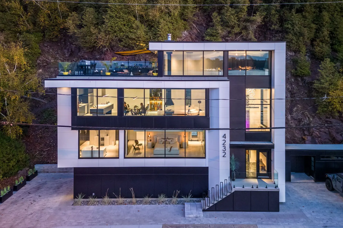 Modern three-story house with black and white exterior, large windows, and rooftop patio. Trees in background.