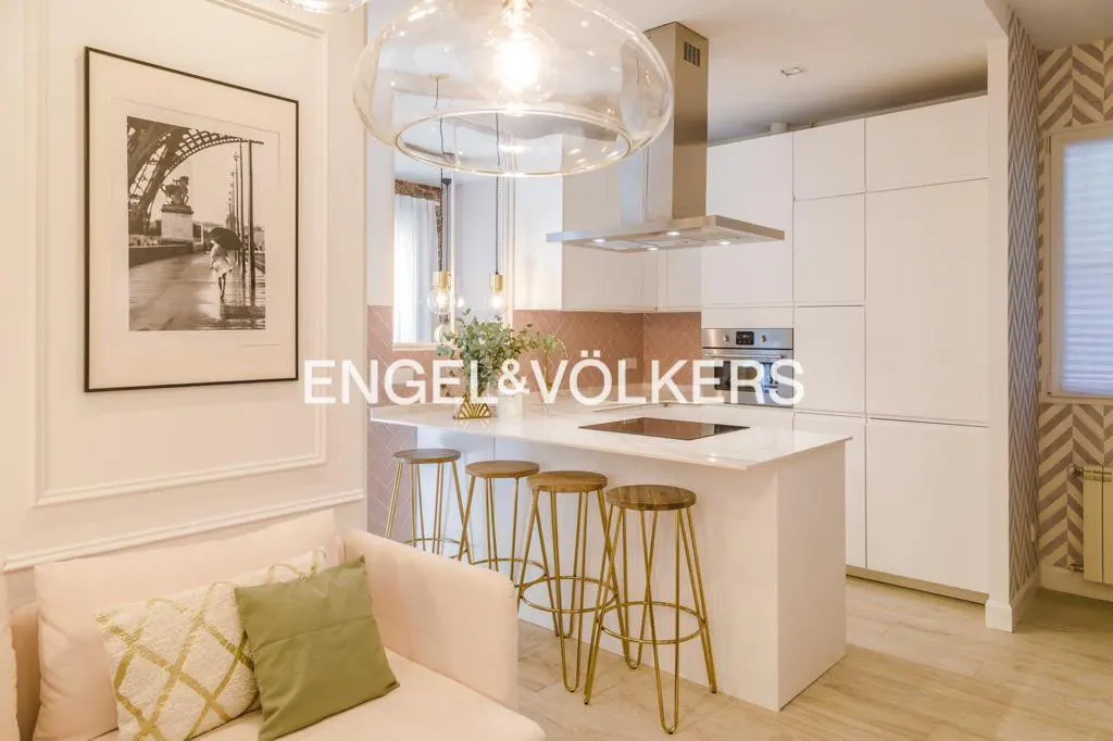 Interior view of a modern kitchen with white cabinets, a marble countertop island with gold stools, and a pink sofa. A black and white Eiffel Tower print hangs on the wall.