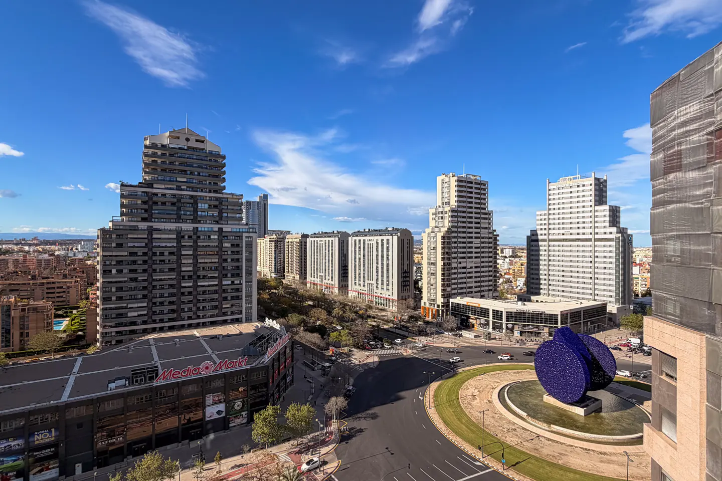Cityscape view with modern buildings under a blue sky with clouds. A roundabout with a blue sculpture is in the foreground.