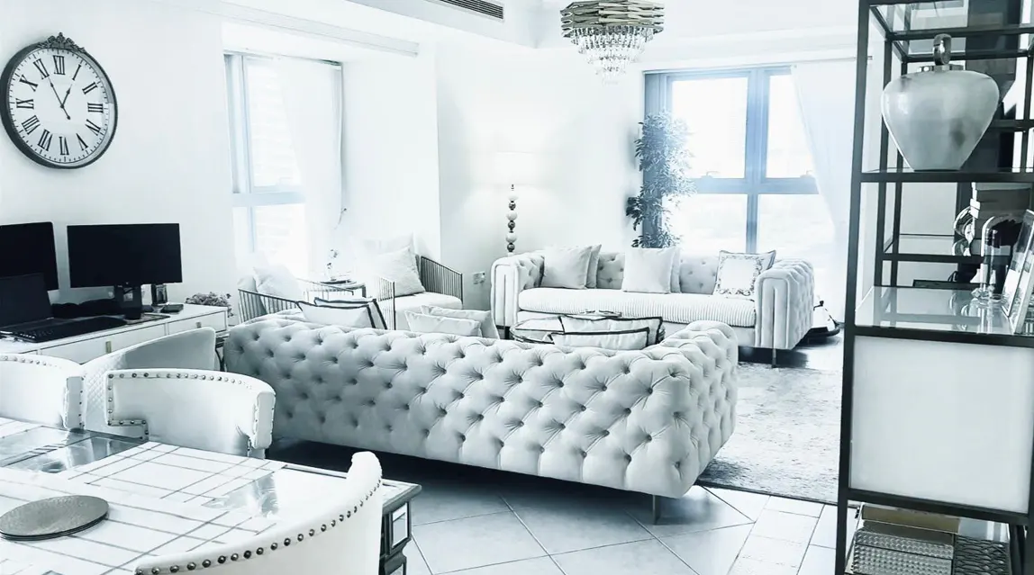 Bright, modern living room with white tufted sofas, chairs, and a glass dining table. A large clock hangs above a desk with monitors.
